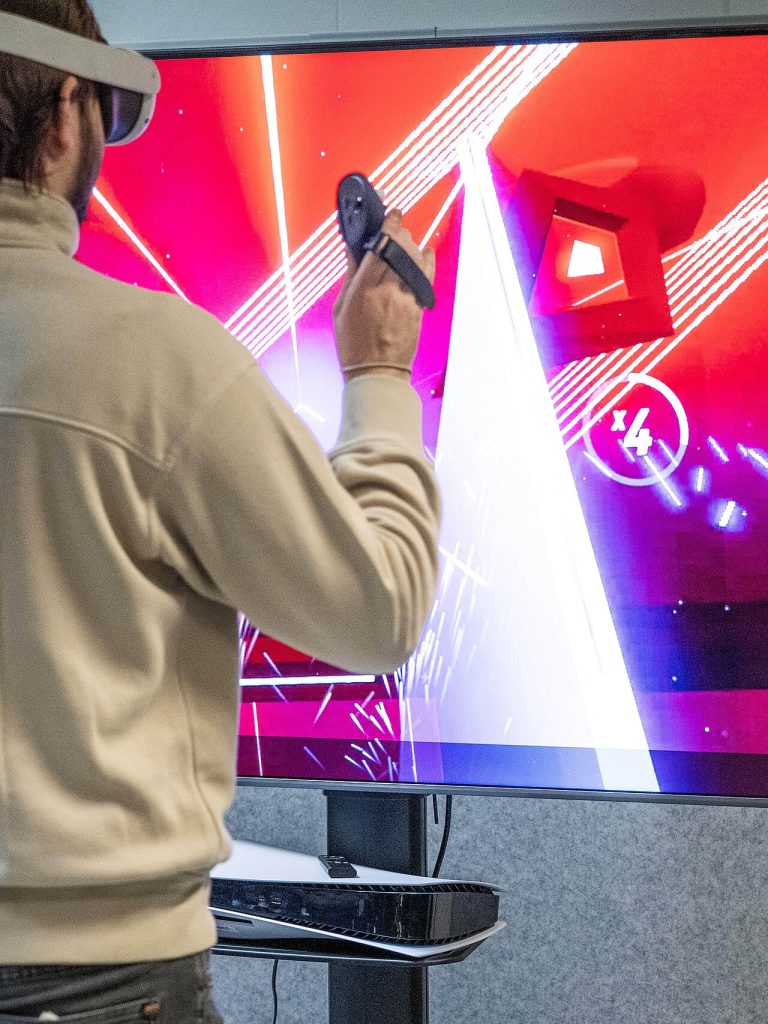 Virtuix Bundles Omni One VR Treadmill With Meta Quest A gamer wearing a virtual reality headset in front of a large immersive display